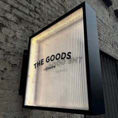 the goods store sign is lit up against a brick wall and has black lettering on it