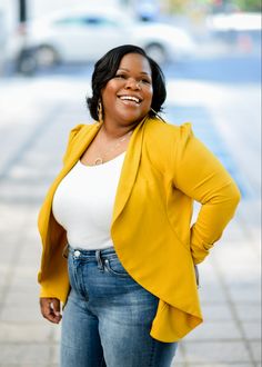 a woman in jeans and a yellow jacket smiles at the camera while standing on a sidewalk