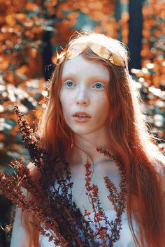 a woman with red hair and blue eyes is standing in front of some leaves, looking at the camera