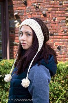 a woman wearing a crocheted hat with pom poms in front of a brick building