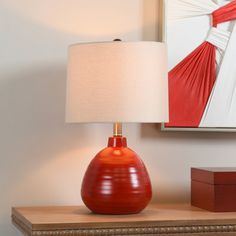 a red vase sitting on top of a wooden table next to a lamp and painting