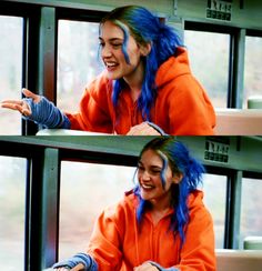 a woman with blue hair sitting in front of a window wearing an orange hoodie