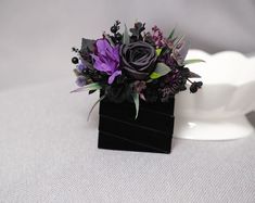 a black vase with purple flowers and greenery in it on a white table cloth