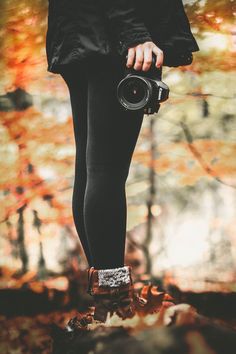 a woman holding a camera in her right hand while standing on top of a leaf covered ground