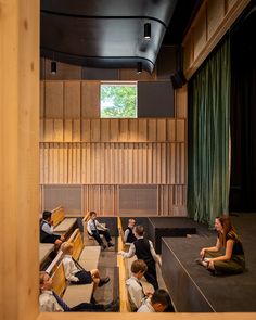 people sitting on benches in an auditorium with green curtained walls and wooden paneling