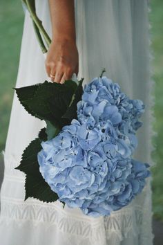 a woman holding a bouquet of blue hydrangea flowers in her hand and wearing a white dress