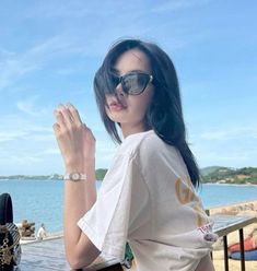 a woman wearing sunglasses sitting at a table next to the ocean with her hand on her hip