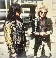 two young men standing next to each other in front of a telephone booth, one wearing sunglasses and the other with long hair