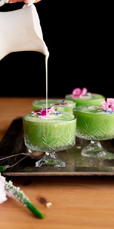 a person pouring white liquid into small green desserts on a tray with flowers and greenery