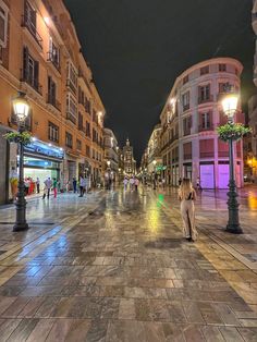 a woman walking down the middle of a city street at night with buildings in the background