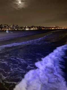 the beach is covered in blue water and waves at night with city lights in the background