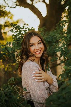 a woman is smiling and posing for the camera with her arms around her chest, surrounded by greenery