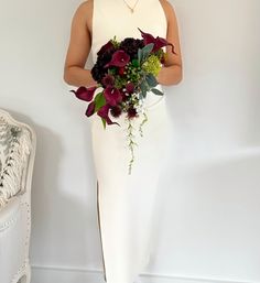 a woman in a white dress holding a bouquet of red and green flowers, standing next to a chair