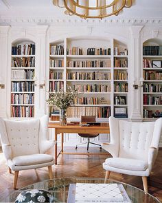 two white chairs sitting in front of a wooden table and bookshelf filled with lots of books