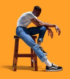 a man sitting on top of a wooden stool next to an orange background with his legs crossed