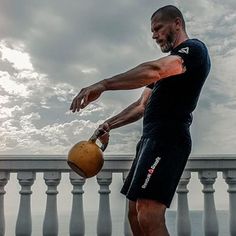 a man holding a kettle while standing next to a railing with clouds in the background