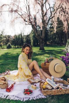 a woman sitting on a blanket in the grass with picnic foods and drinks around her