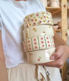 a woman holding three cups with flowers on them in front of shelves and she is wearing white t - shirt