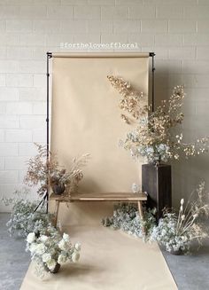 an arrangement of white flowers and greenery in front of a beige backdrop with a wooden bench