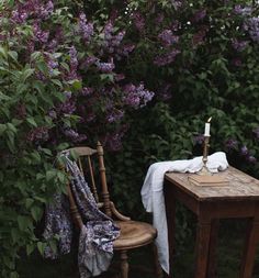 a table and chair with a candle on it in front of purple flowers near a bush