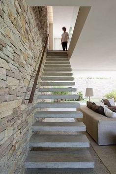 there is a woman walking down the stairs in this modern house with stone walls and floor to ceiling windows