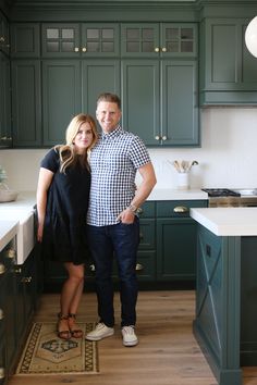 a man and woman standing in the middle of a kitchen with dark green cabinetry