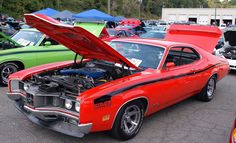 an orange muscle car with its hood open in a parking lot next to other cars