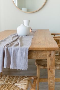 a wooden table topped with a white vase next to a mirror and wicker chairs