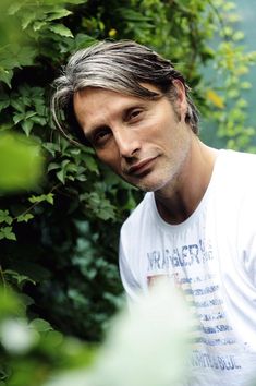 a man with grey hair standing in front of some bushes and trees looking at the camera