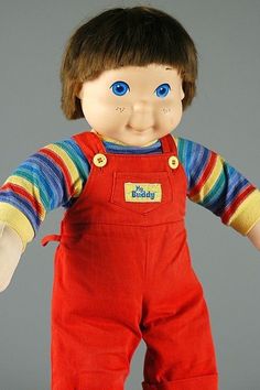 a doll with blue eyes and red overalls is posed in front of a gray background