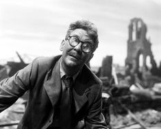 black and white photograph of man in suit with destroyed buildings behind him, looking at camera