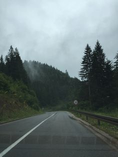 an empty road in the mountains with trees on both sides and foggy skies above