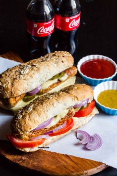 two sub sandwiches sitting on top of a wooden cutting board next to bottles of soda