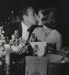 an old black and white photo of two people kissing at a table with flowers on it