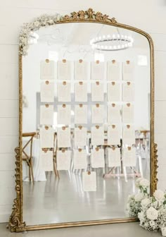 a gold framed mirror with seating cards on it and flowers in front of the mirror