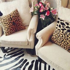 two white chairs with leopard print pillows and pink flowers in a vase next to them