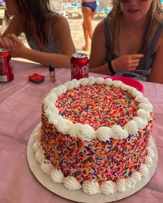 two women sitting at a table with a cake and sodas in front of them