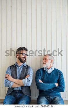 two men sitting next to each other in front of a white wall with their arms crossed