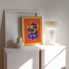 an orange and blue art piece sitting on top of a white cabinet next to a vase