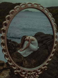 a woman sitting on top of a rock next to the ocean in front of a mirror