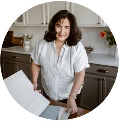 a woman standing in front of a kitchen counter holding an open book and smiling at the camera