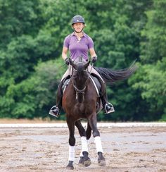 a woman riding on the back of a brown horse across a dirt field with trees in the background