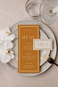 a table setting with an orange menu card and white orchids on the place cards