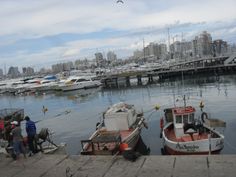 several small boats docked in the water near a dock with people standing on it and buildings in the background