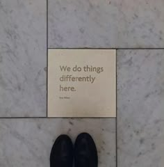 there is a sign that says we do things differently here on the marble floor next to a pair of black shoes