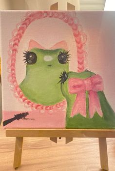 a painting of a green cat wearing a pink bow and sitting on a wooden easel