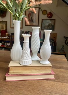 three white vases sitting on top of a wooden table next to books and flowers