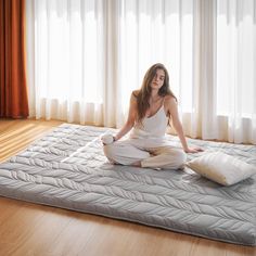 a woman sitting on top of a mattress in front of a window with white curtains