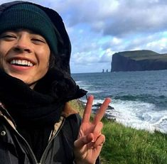 a woman standing on top of a lush green field next to the ocean giving the peace sign