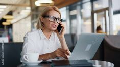 a woman sitting at a table with a laptop and talking on the phone while holding a coffee cup